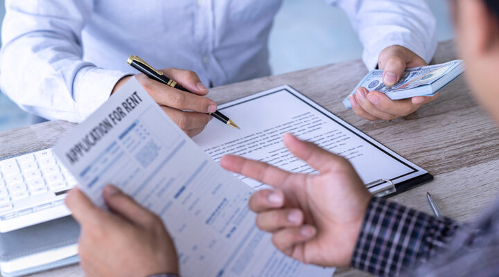Two individuals at a table are engaged in a transaction. One person is holding paperwork labeled "APPLICATION FOR RENT" and pointing to it, while the other is handing over cash and holding a pen, indicating a rental agreement process. A keyboard is visible in the background.