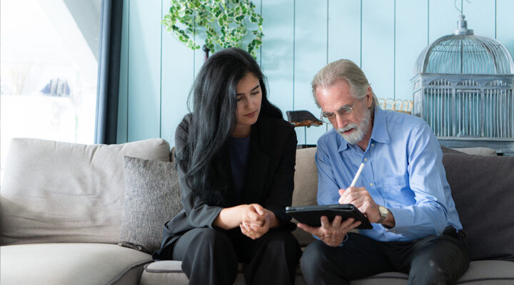 A woman with long black hair and a man with grey hair and glasses sit on a couch, looking at a tablet. The man is using a stylus to write on the tablet while the woman attentively watches. The background has light blue walls, a birdcage, and a potted plant.