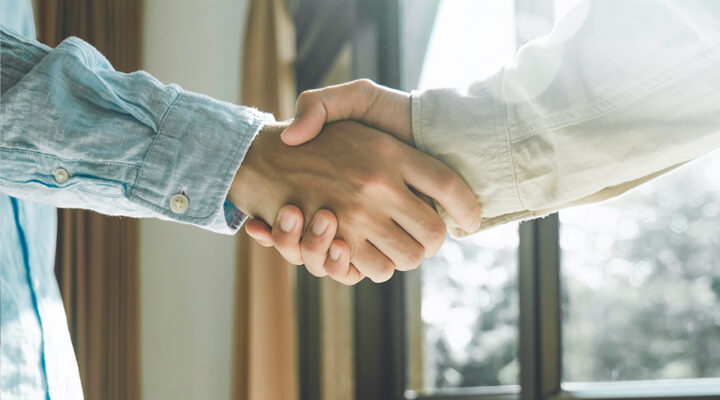 Two people shaking hands indoors near a window with sunlight streaming in. One person wears a denim shirt, and the other a light-colored jacket. The image conveys a sense of agreement or partnership.