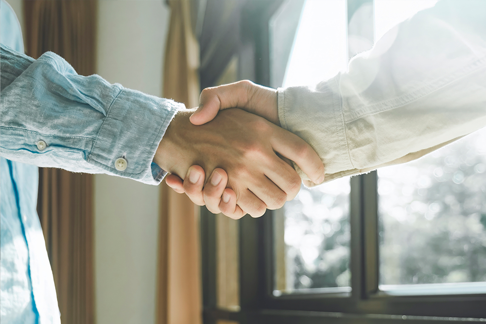 Two people shaking hands indoors near a window with sunlight streaming in. One person wears a denim shirt, and the other a light-colored jacket. The image conveys a sense of agreement or partnership.