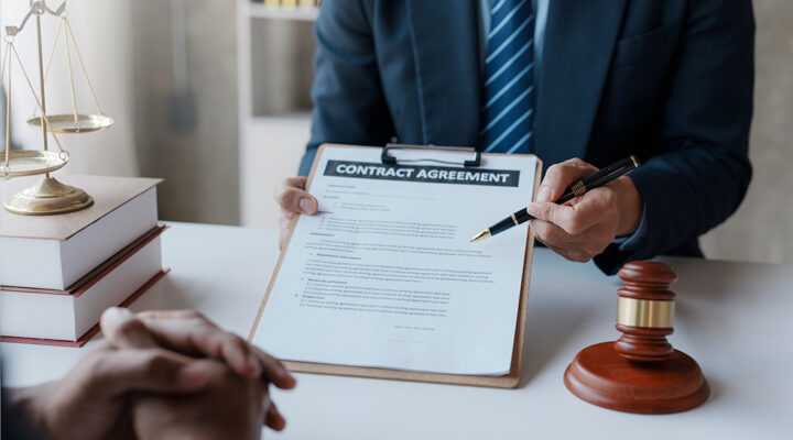 A person in a suit holds a clipboard with a contract agreement in front of another person. The scene includes a gavel, a stack of books, and a set of scales, suggesting a legal or business setting. The person is pointing at the document, indicating discussion or review.