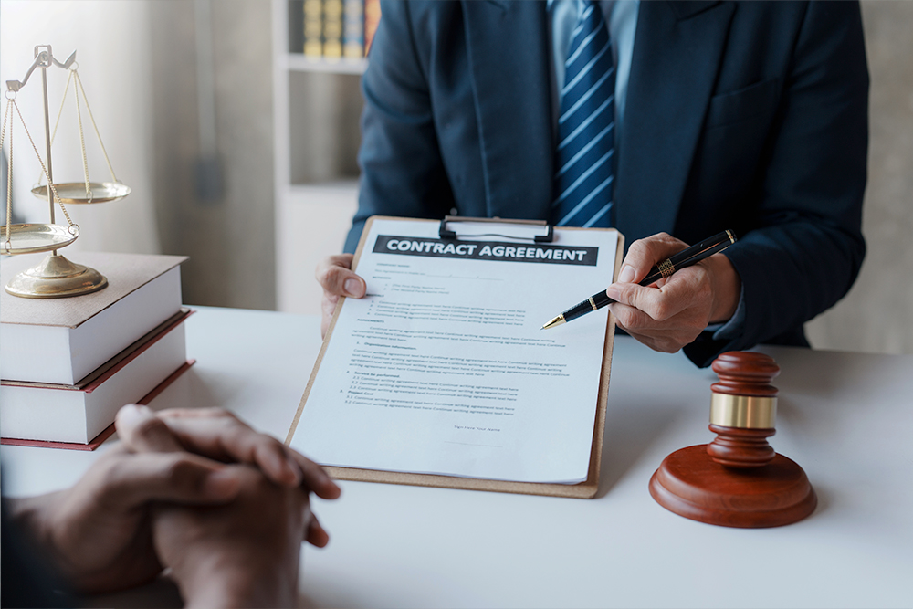 A person in a suit holds a clipboard with a contract agreement in front of another person. The scene includes a gavel, a stack of books, and a set of scales, suggesting a legal or business setting. The person is pointing at the document, indicating discussion or review.