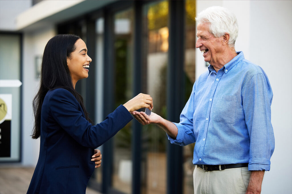 A young woman with long dark hair and dressed in a dark blazer hands over keys to an older man with short white hair wearing a light blue shirt. Both are smiling and standing outside a building with large windows.